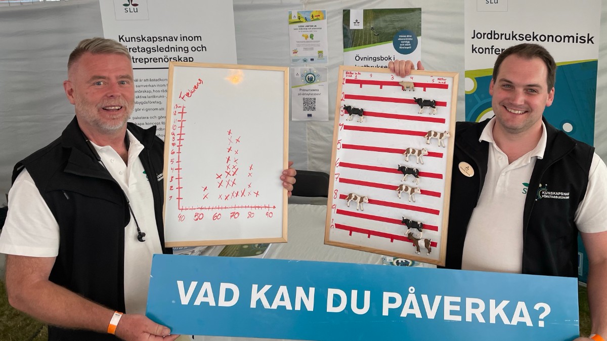 Två män i vita tröjor och svarta västar som håller i blå skylt med vit text.
