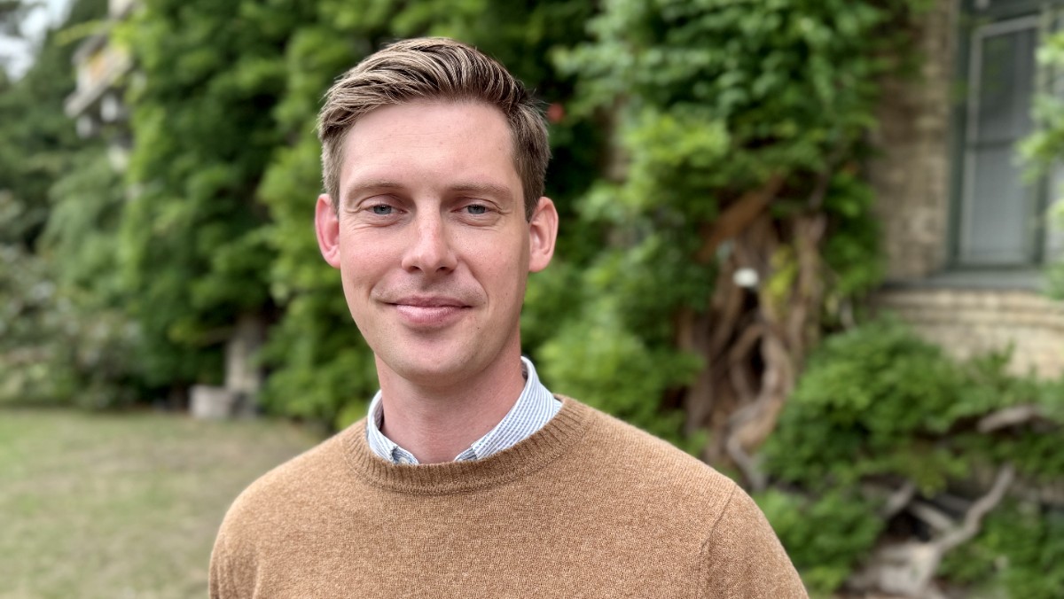 Man med kort hår och brun tröja mot grönskande bakgrund